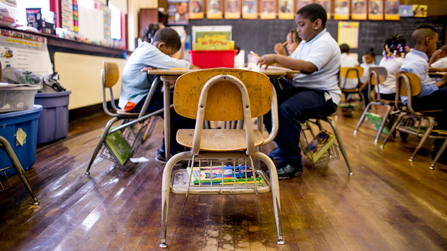 Students sit at tables in a classroom with a wooden floor an a window on the left and a chalkboard in the background.