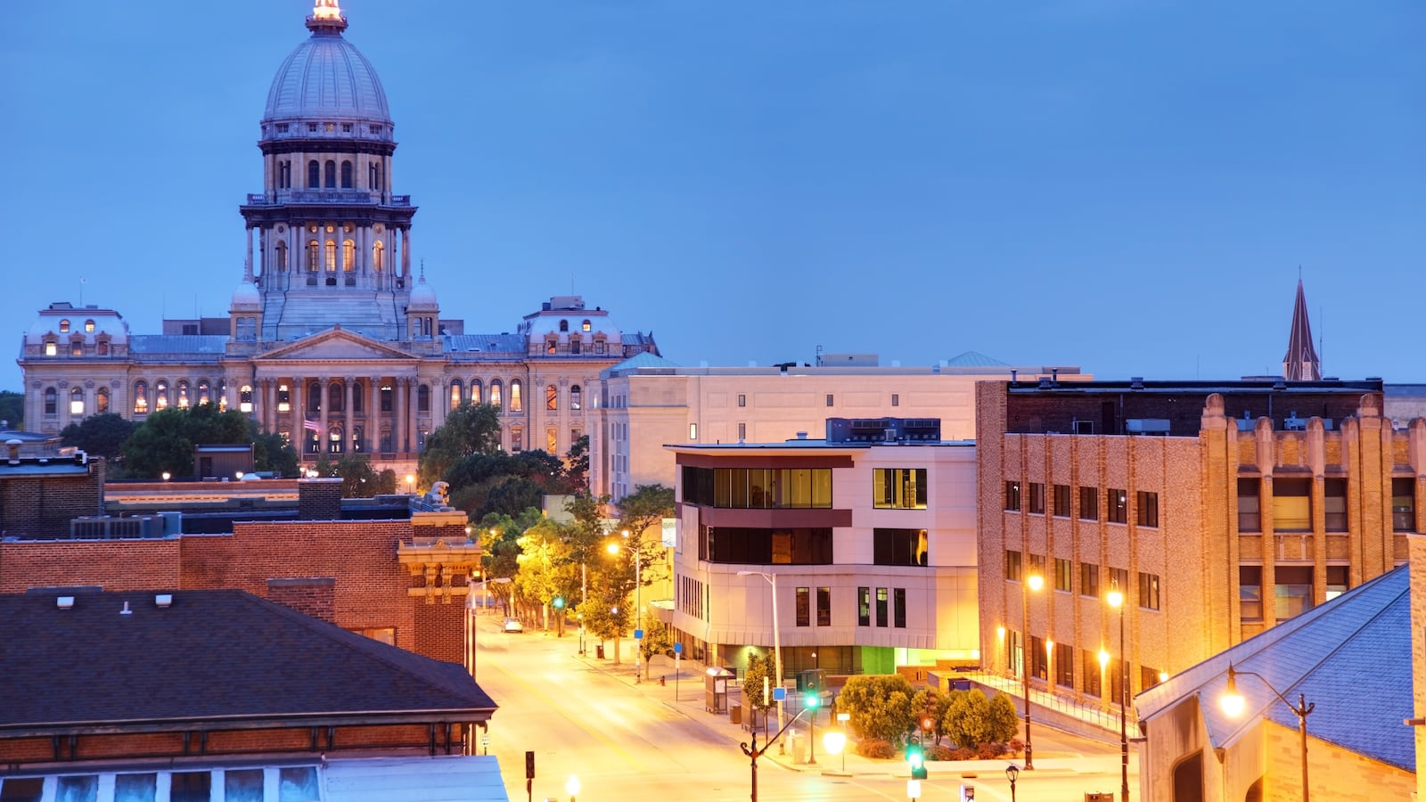 The Illinois State Capitol is seen along with other buildings and a street illuminated during the early evening.