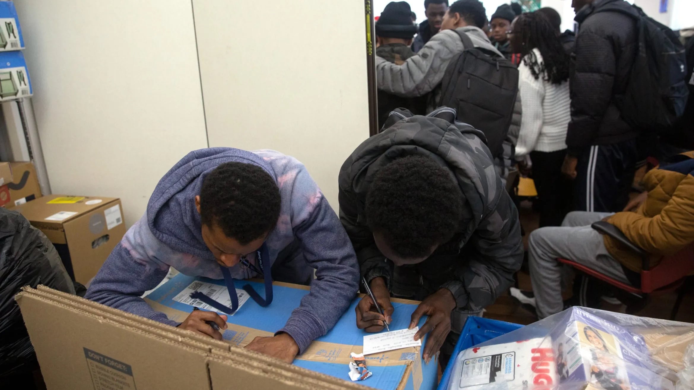 Two young people are huddled over a desk filling out paperwork, while another group is standing in the background.