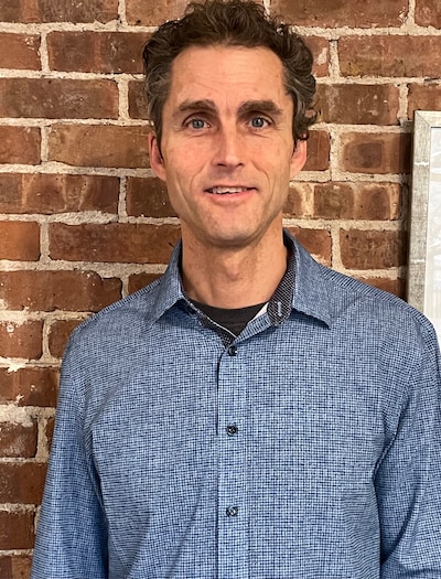 Photo of a man with dark hair. He is wearing a blue button-down shirt and stands against a brick backdrop.