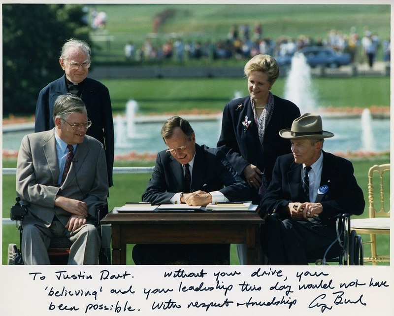 A photo of President George H. W. Bush signing the Americans with Disabilities Act, surrounded by those who advocated for the bill. Below the photo is an inscription thanking Justin Dart that reads, “Without your drive, your ‘believing’ and your leadership this day would not have been possible. With respect and friendship, George Bush.”