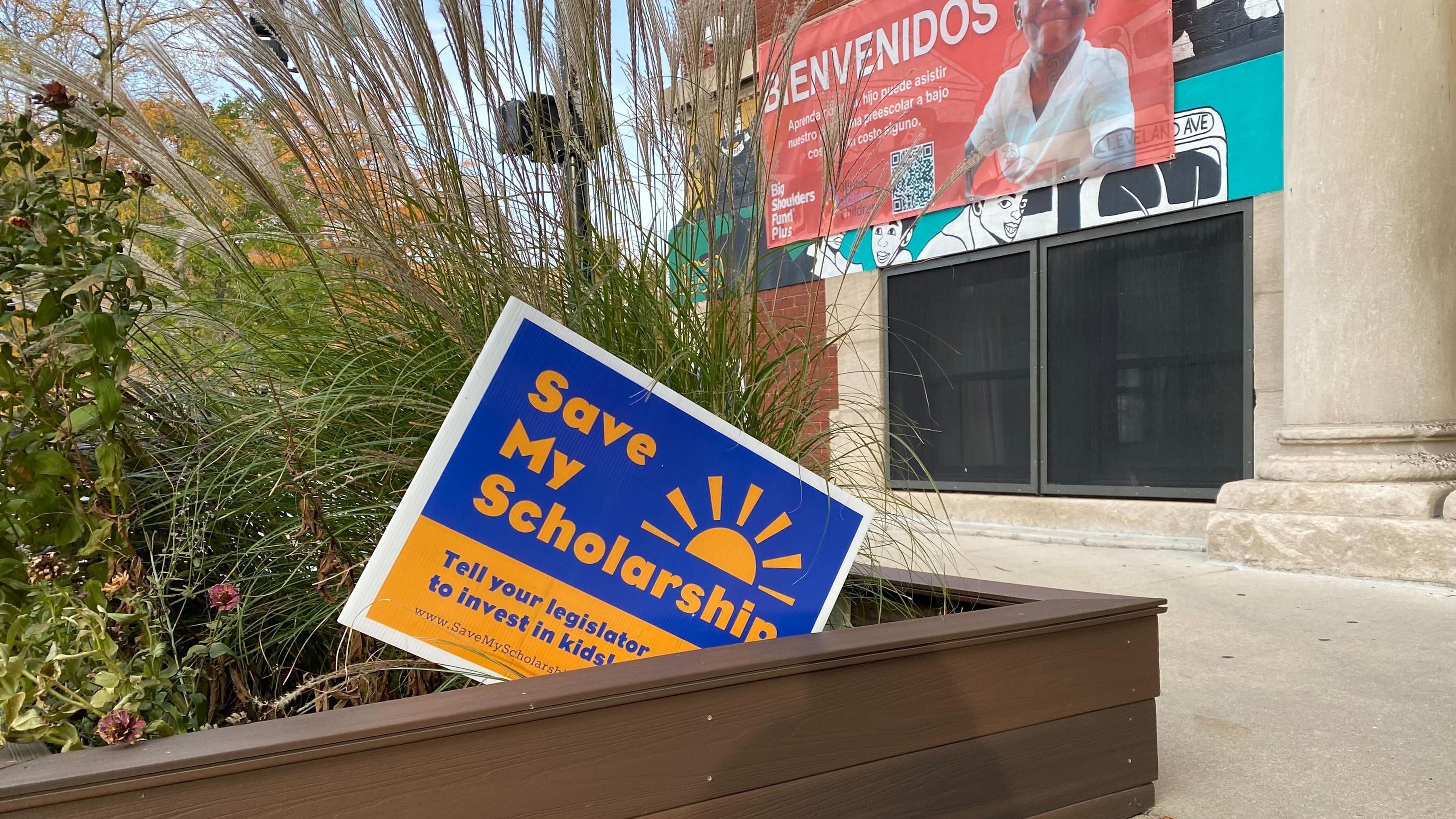 A blue and yellow sign that reads" save my scholarship" sits in a flower bed in front of a school building.