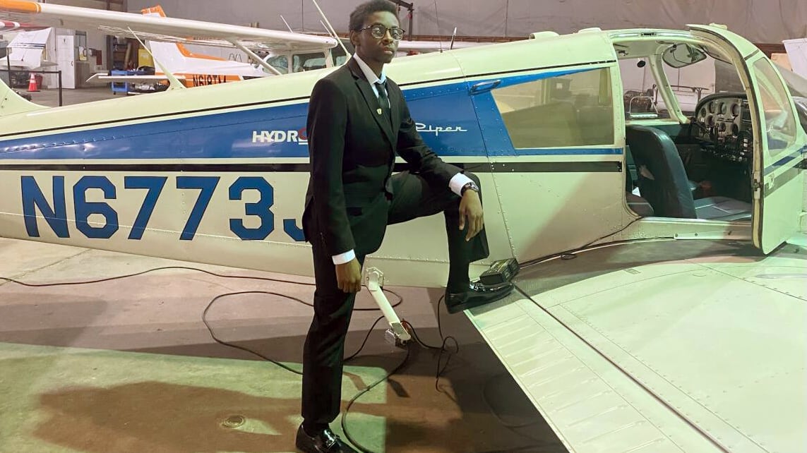 A high school student wearing a dark suit poses by the wing of a plane with a warehouse in the background.