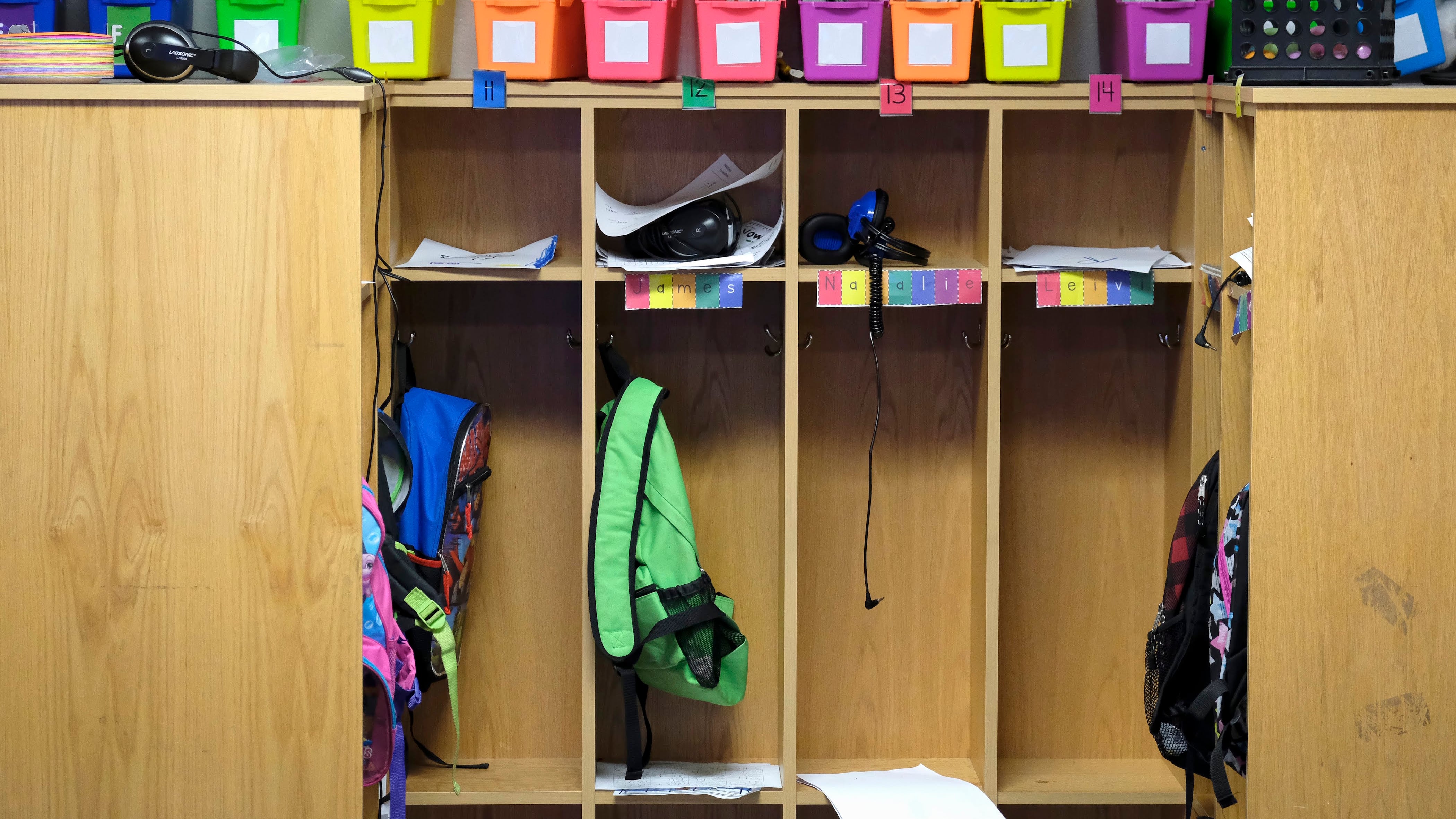 A set of cubbies in a classroom with a couple of backpacks hanging and a row of colorful organizers at the top.