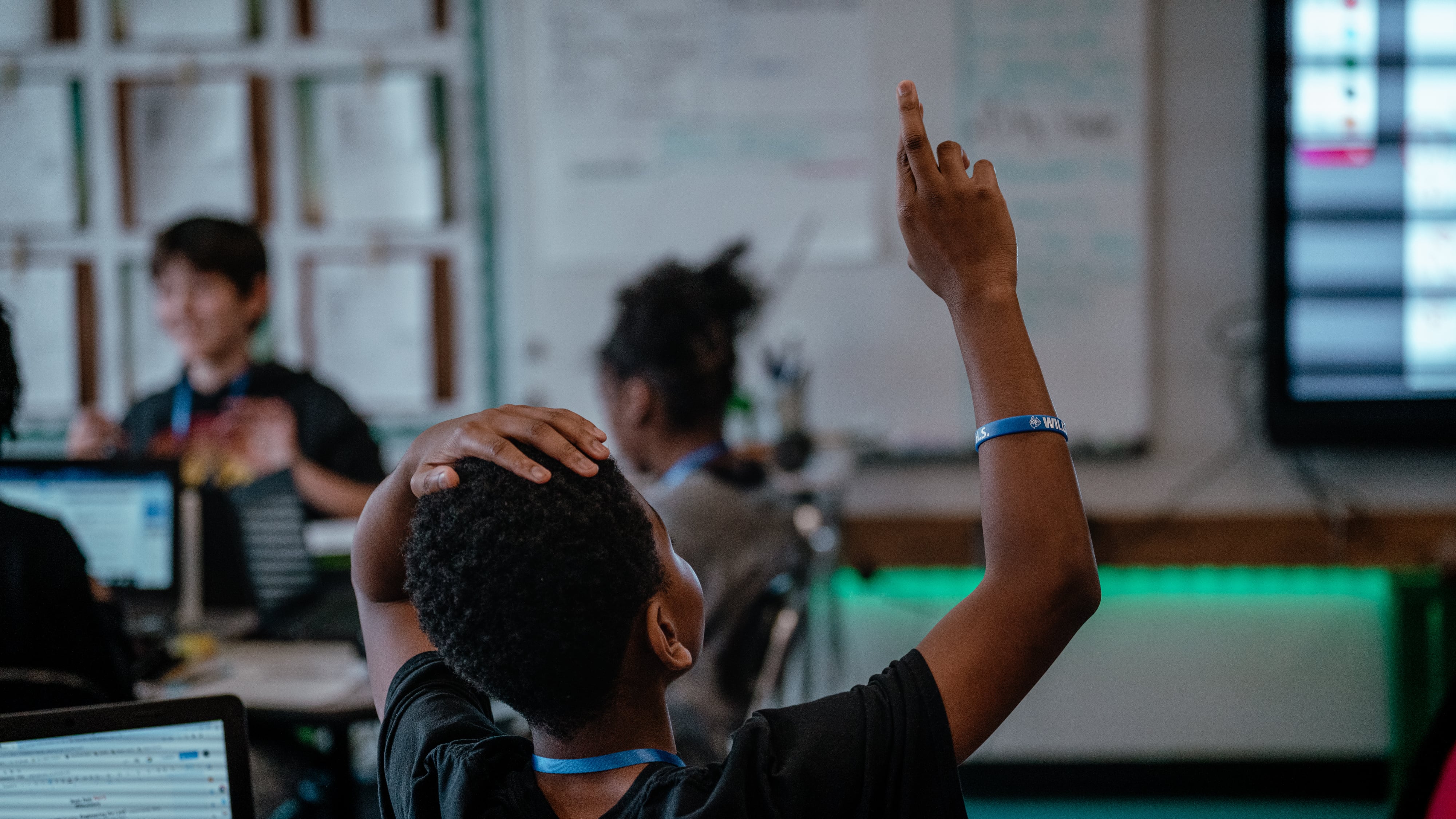 A back view of a student raising their hand in a classroom with other students in the background.