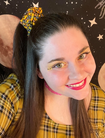 Young woman with brown hair and green eyes wears a yellow plaid top and a matching hair tie.