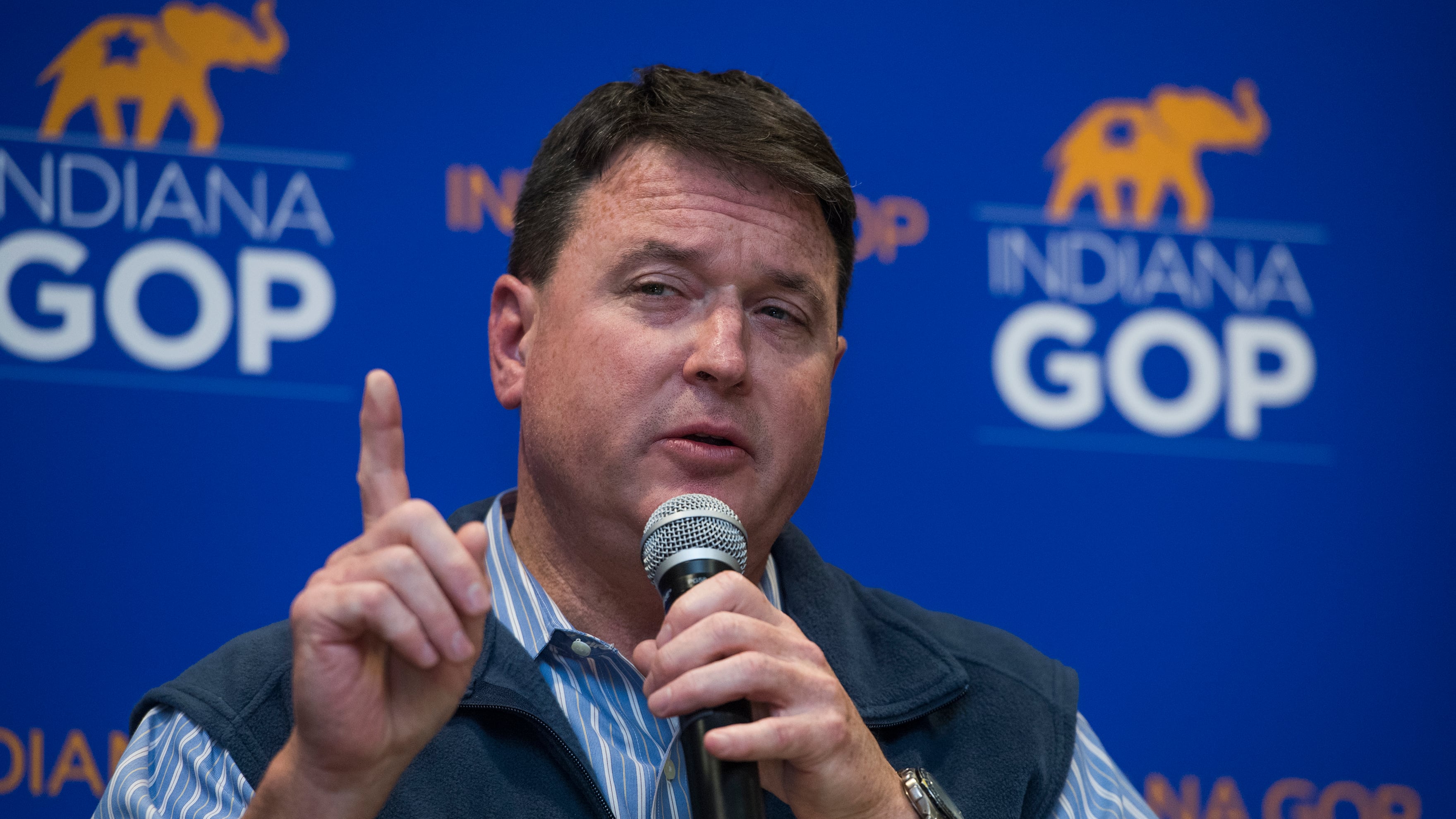 A man with short dark hair and wearing a dark vest over a blue and white stripe shirt holds a microphone and points with his hand. There is a blue background with small elephants and logos that read "Indiana GOP."