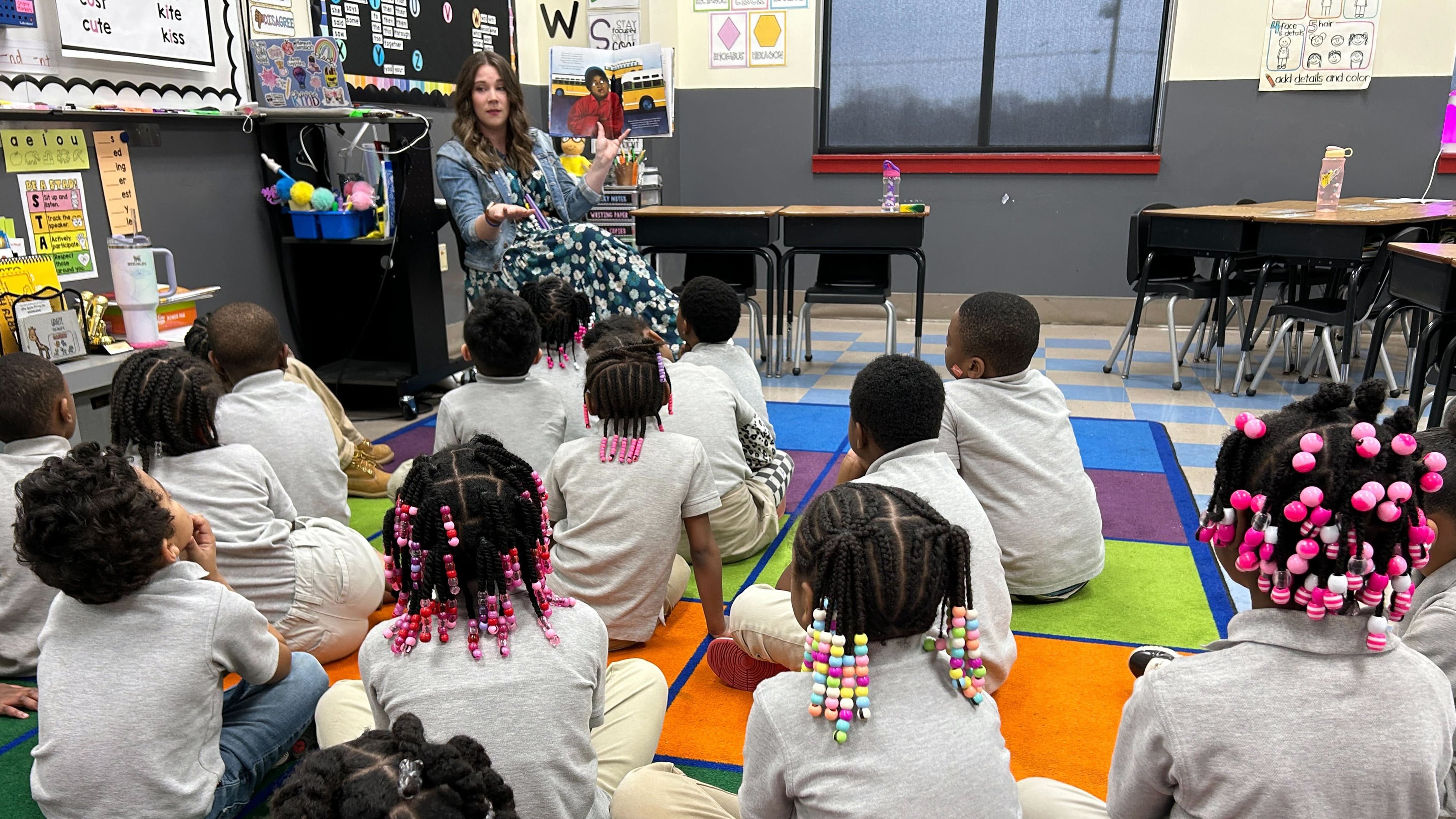 A teacher with long brown hair holds up a picture book to a class full of young students sitting on a colorful rug in a classroom. Students are wearing a grey shirt and light tan pants as a uniform and many students have colorful beads in their braids.