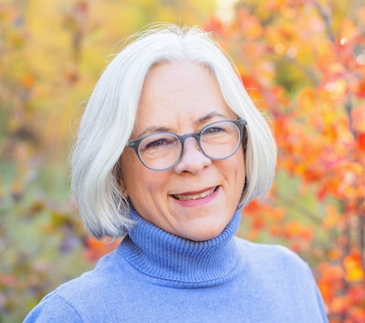 Headshot of a white woman with white hair. She wears a lilac sweater. Fall folliage is behind her.