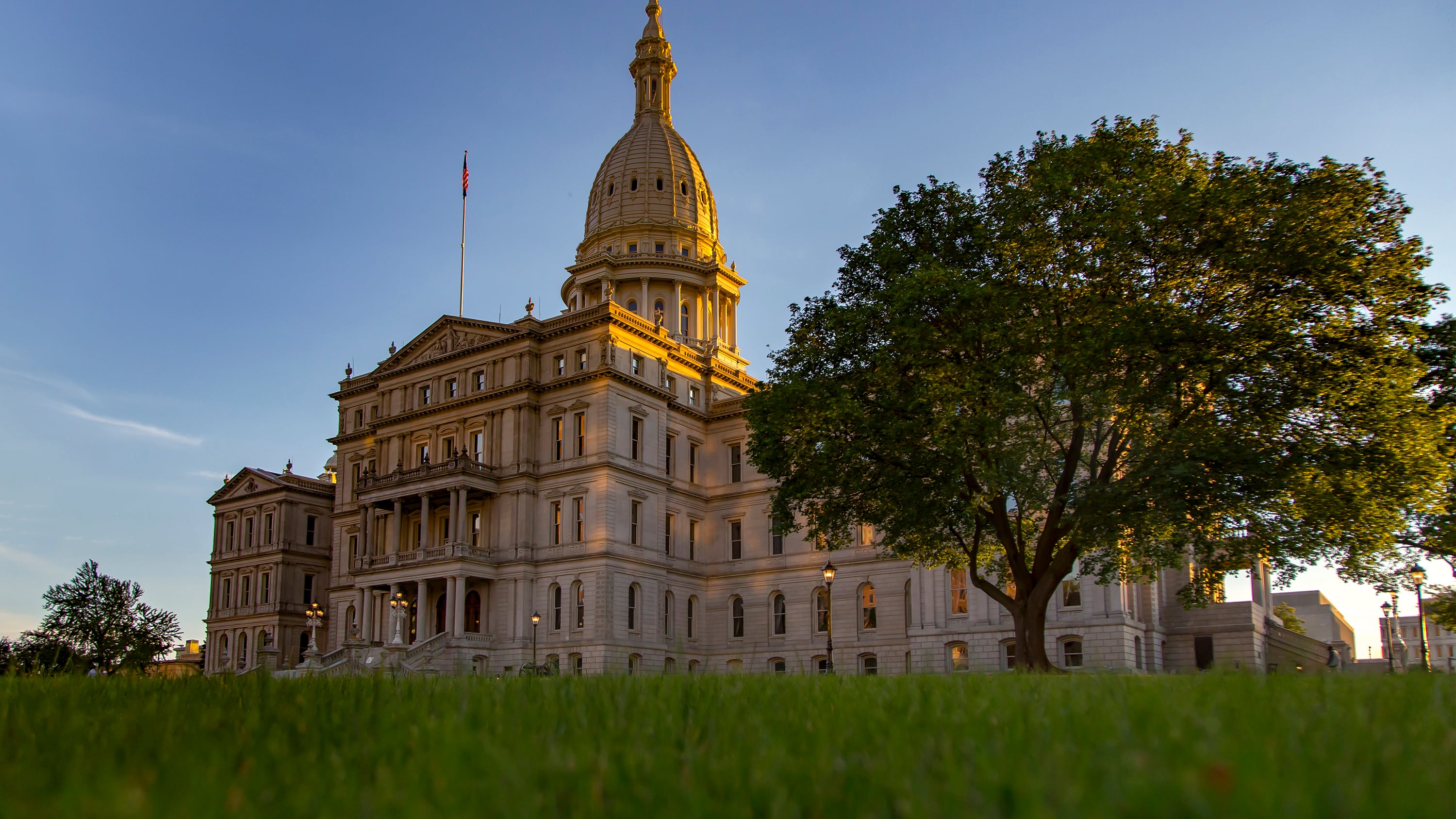 The sun is setting on the capitol building with a large green tree in the foreground and blue sky in the background.