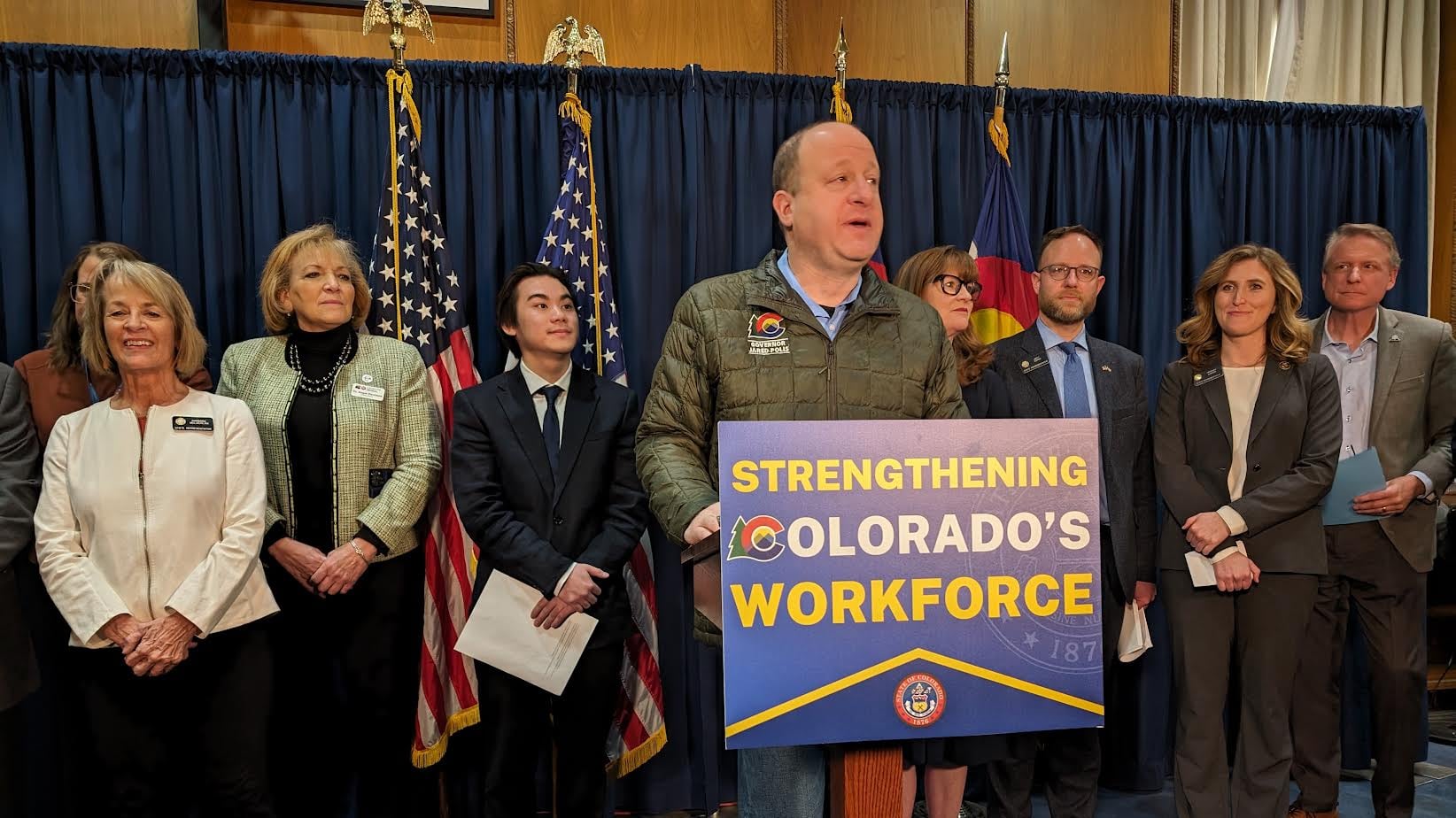 A man wearing a green jacket stands at a podium with a sign that reads "Strengthening Colorado's workforce." There is a group of people standing in a row with flags in the background.