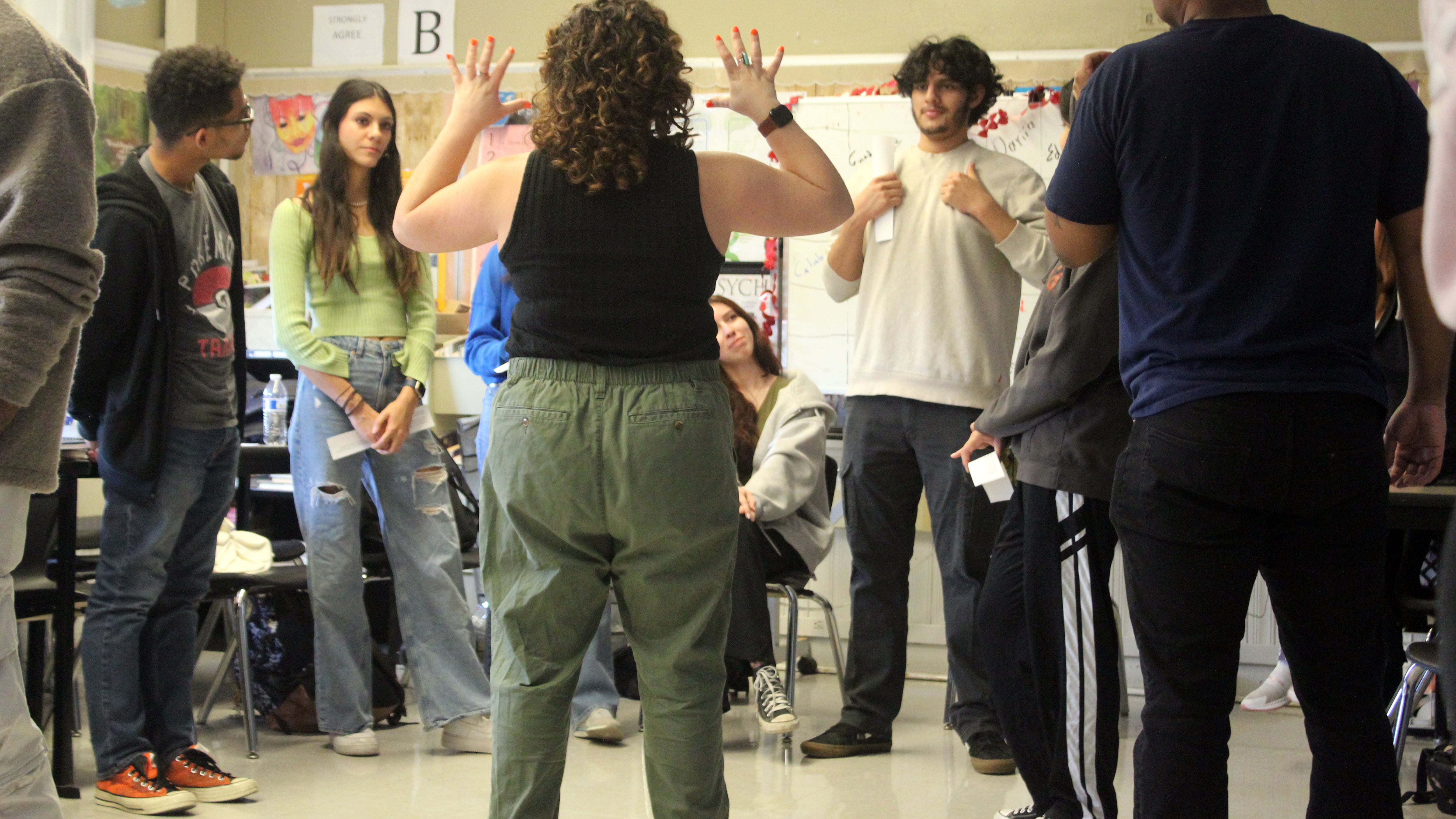 An adult holds her hands up in front of a group of high school students standing in a classroom.