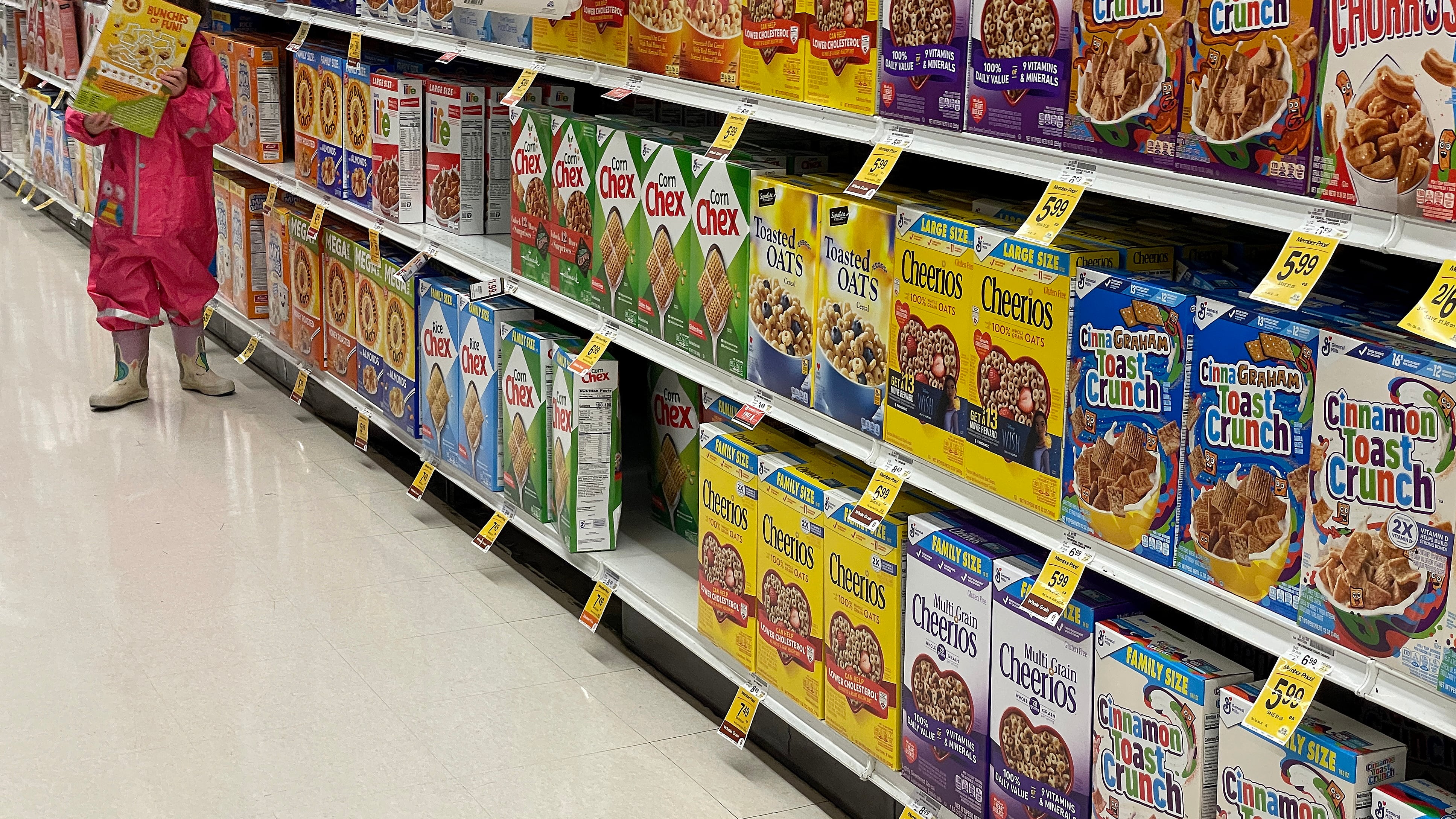 A young child carries a large box of cereal covering their face while they stand next to an aisle full of cereal boxes.