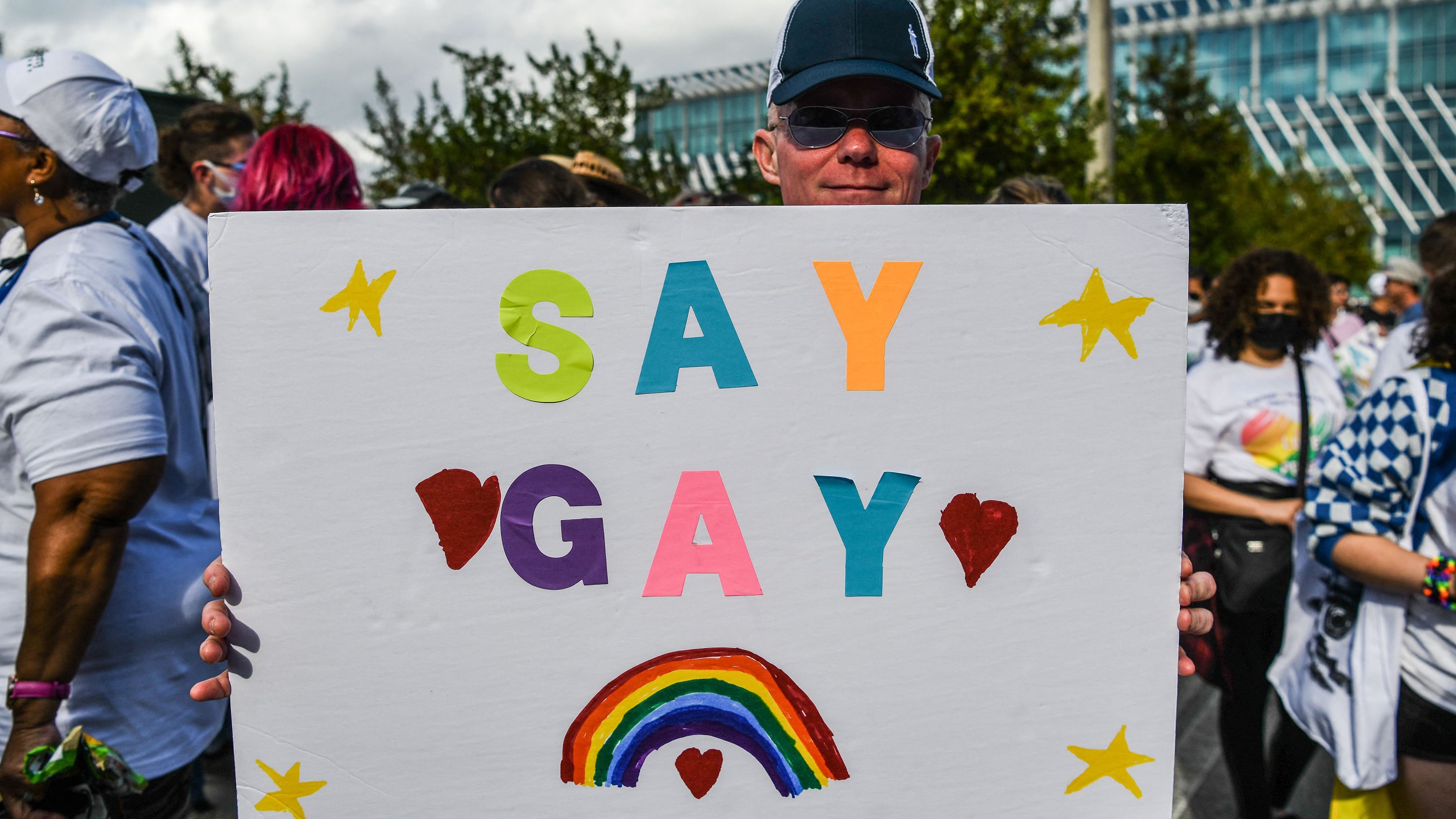 A person wearing a hat and holding a giant white sign with colorful letters that say "SAY GAY" with a rainbow, stars and hearts with people standing in the background.