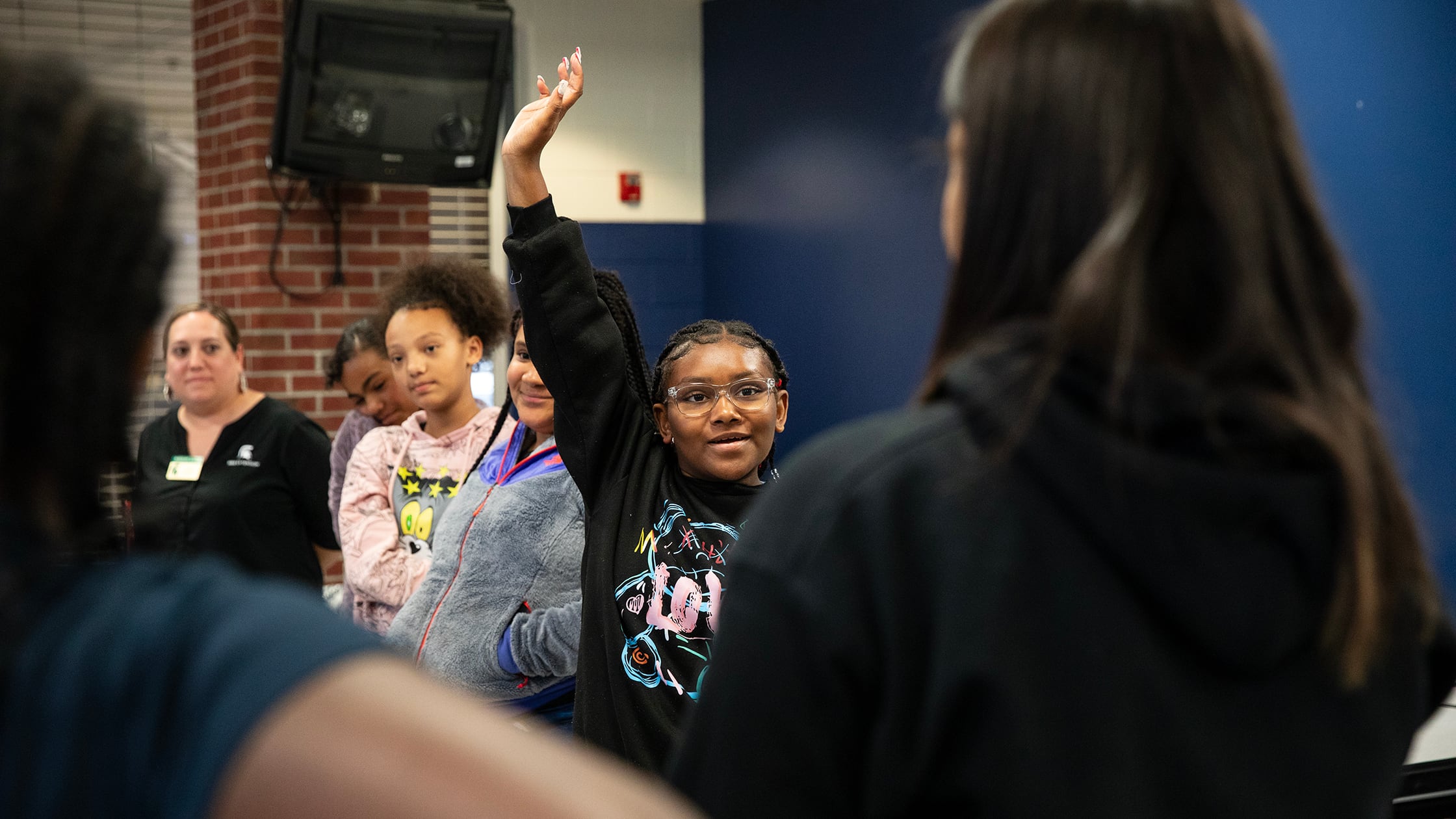 A student wearing a black sweater holds her hand up in a circle of other students and adults. There is a dark blue wall and a red brick column in the background.