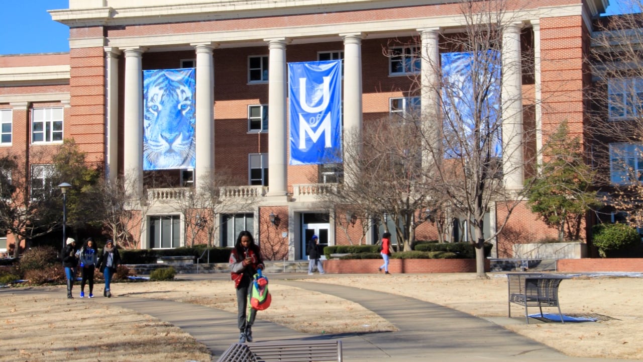 Students walk along a sidewalk in front of a building on The University of Memphis campus.